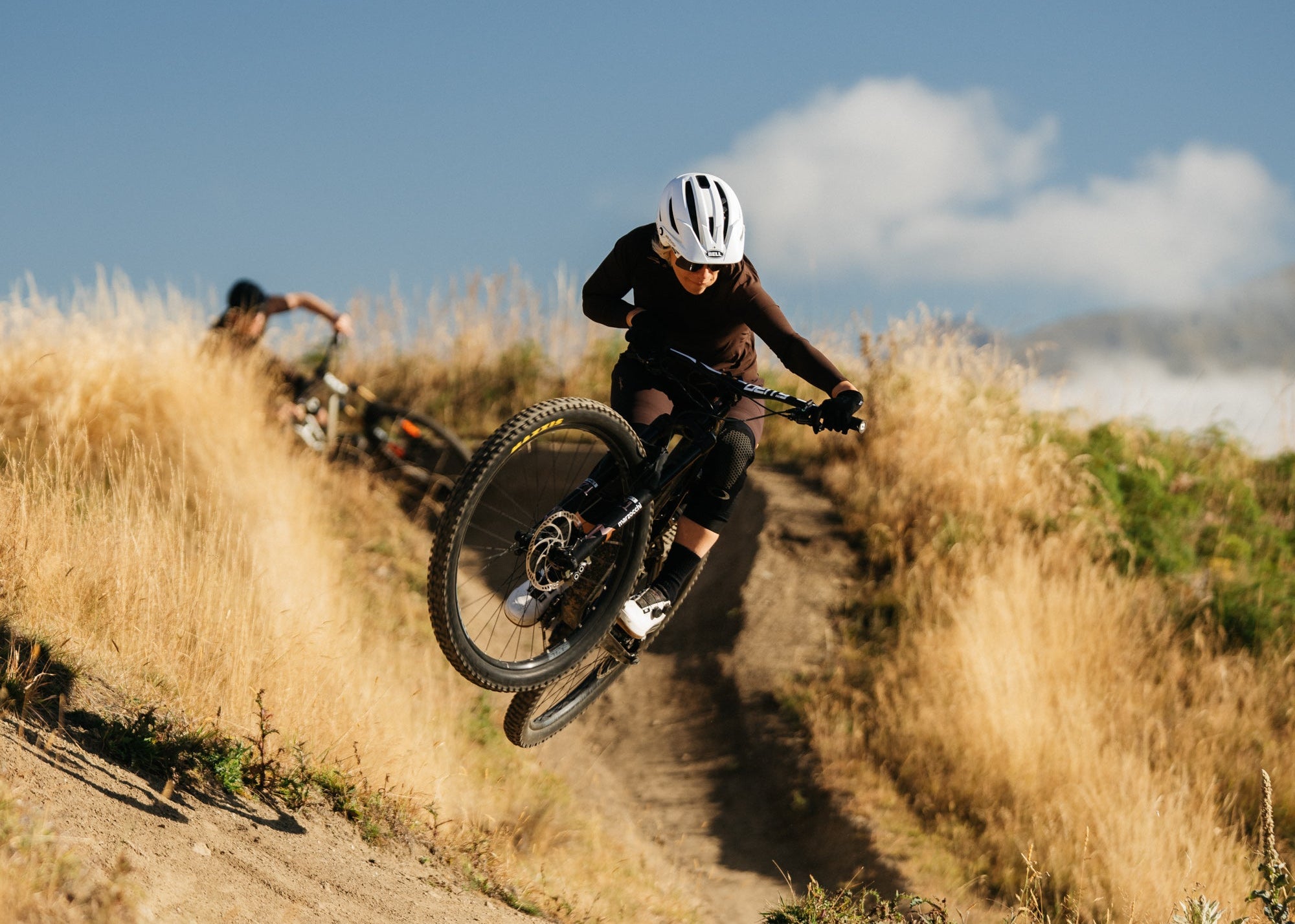Two mountain bikers riding on a dirt path with a clear blue sky and clouds in the background.