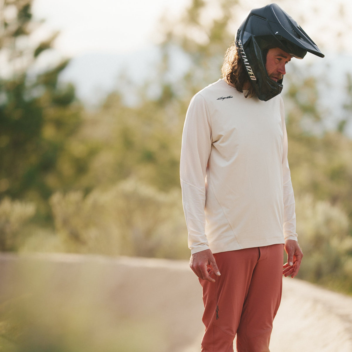 Matt Miles wearing a helmet and white long-sleeve shirt standing on a dirt path with trees in the background